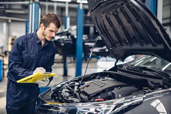 Mechanik w granatowym kombinezonie notuje coś na żółtej teczce, stojąc przy otwartej masce samochodu. To skrót do świata koni mechanicznych.