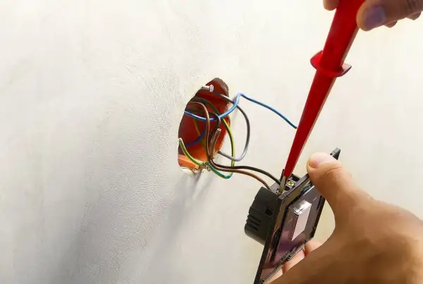 Instalator podłącza podwójny włącznik światła 2 kable, używając próbnika napięcia. Widać przewody w puszce instalacyjnej.
